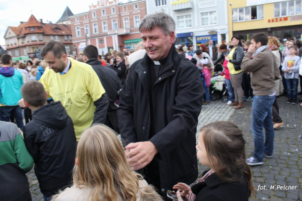 Mężczyzna w czarnym płaszczu rozmawia z dziećmi na tle tłumu i budynków.