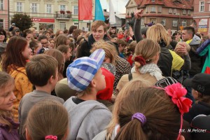 Tłum ludzi zgromadzony na placu miejskim, w tle budynki i flagi.