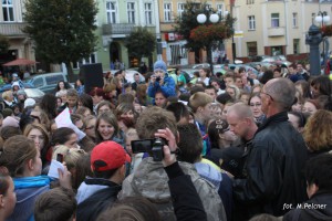 Tłum ludzi zgromadzony na placu miejskim, w tle budynki i lampy.