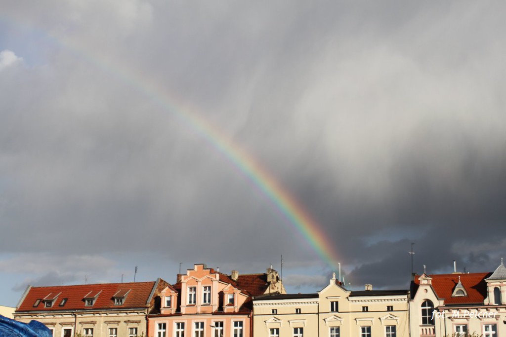 Tęcza nad historyczną zabudową miasta z czerwonymi dachami, w tle zachmurzone niebo.