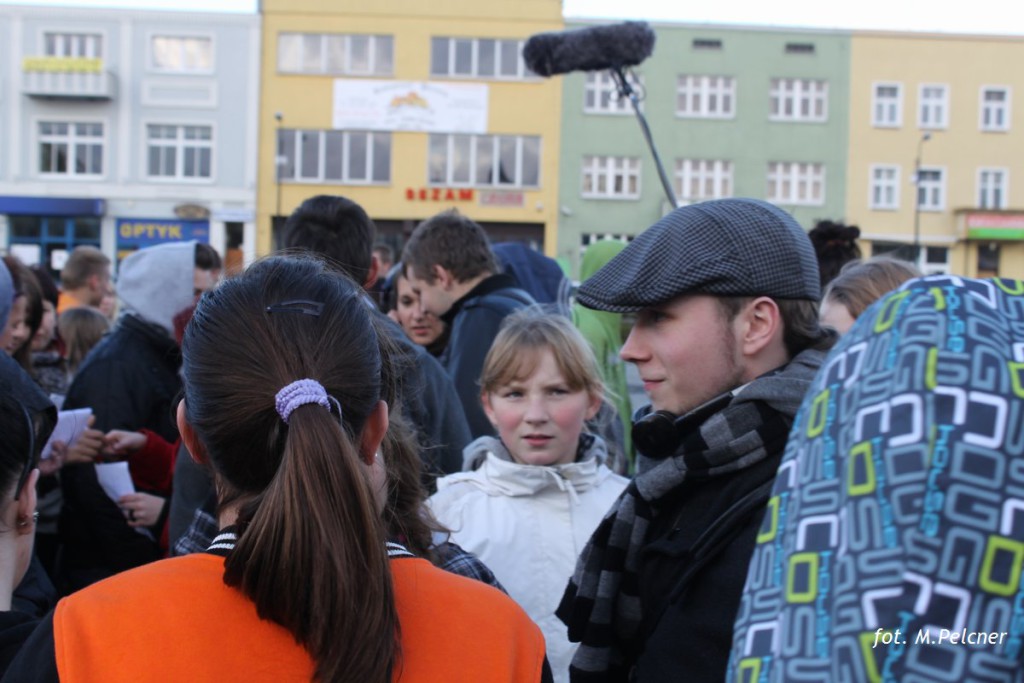 Grupa osób na placu miejskim, nagrywana przez ekipę filmową z mikrofonem.