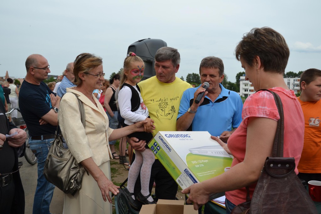 Mężczyzna wręcza paczkę z telewizorem Grundig podczas wydarzenia plenerowego, otoczony tłumem osób.