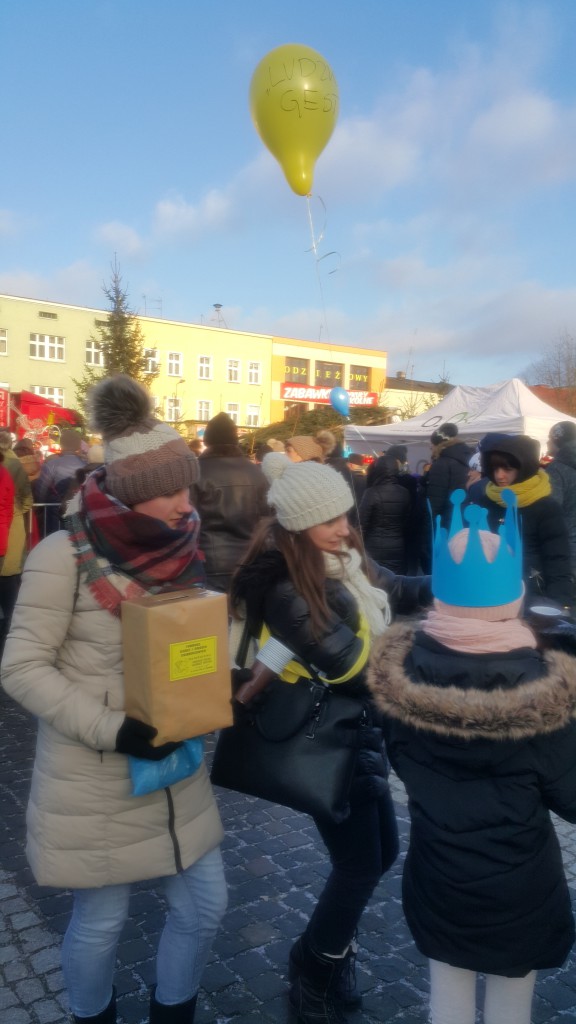 Ludzie uczestniczą w Orszaku Trzech Króli, jeden trzyma pudełko, drugi balon z napisem, w tle budynki i tłum.