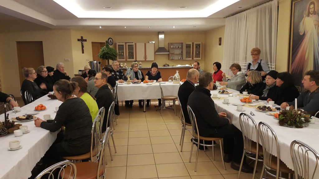 Grupa osób uczestniczy w mszy świętej w pomieszczeniu z długimi stołami nakrytymi białymi obrusami.