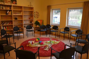 Pokój z krzesłami ustawionymi w krąg, regałem z książkami i oknem z widokiem na zewnątrz.