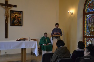 Mężczyzna odczytuje tekst przy stole, w obecności księdza i osób w pomieszczeniu z witrażem.
