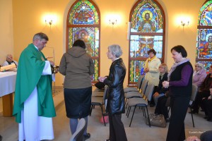 Kapłan odprawia ceremonię eucharystyczną w obecności wiernych w jasnym pomieszczeniu z witrażami.