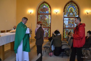 Uroczystość religijna w kaplicy: ksiądz odprawia obrzęd w obecności wiernych, w tle widoczne witraże.
