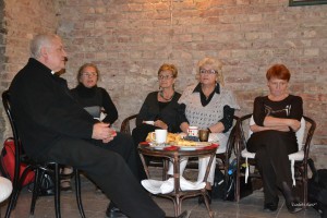 Grupa osób siedzi przy stoliku w pomieszczeniu z ceglanym murem, na stole znajdują się naczynia z jedzeniem i napojami.