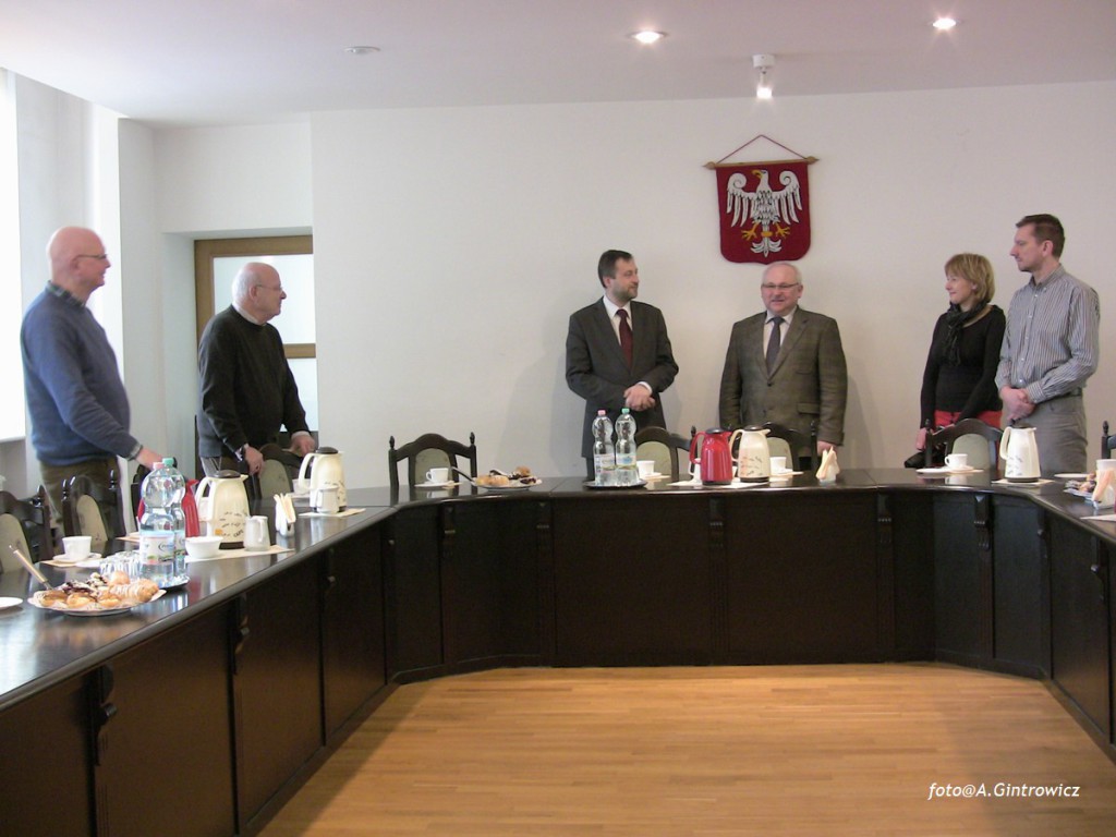 Grupa osób uczestniczy w spotkaniu przy długim stole w pomieszczeniu z herbem na ścianie.