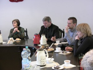 Czterech rozmówców siedzi przy stole, na którym znajdują się napoje, słodkie przekąski i naczynia.