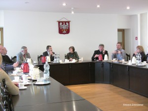 Osoby uczestniczą w spotkaniu przy długim stole w pomieszczeniu z polskim herbem na ścianie.