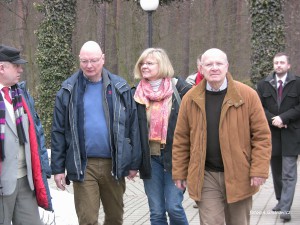 Grupa osób idzie chodnikiem w słoneczny dzień, w tle widoczne drzewa i lampa uliczna.