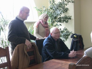 Trzy osoby siedzą przy stole w pomieszczeniu z roślinami, w tle widoczne okno.