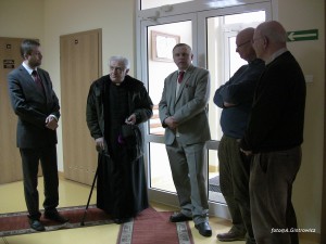 Grupa mężczyzn w garniturach i duchowny rozmawia wewnątrz budynku, widoczne drzwi i wyjście ewakuacyjne.