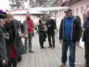 Grupa osób stoi na chodniku przed budynkiem, w tle widać drzewa i samochody.