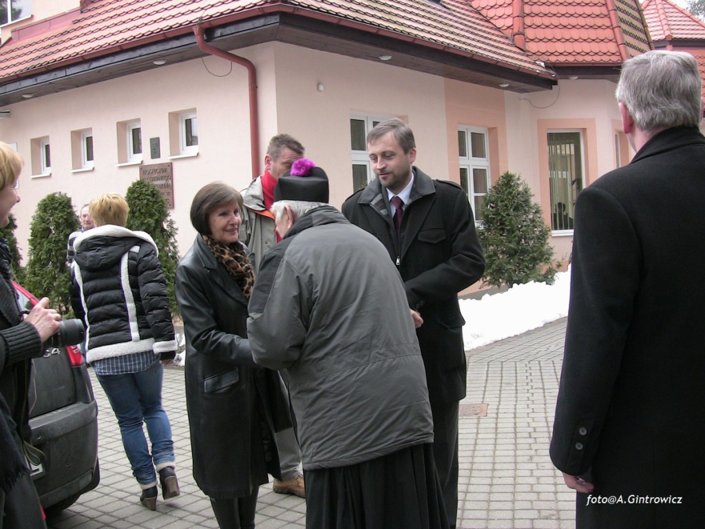 Osoby rozmawiają przed budynkiem z czerwonym dachem, na wyłożonym kostką brukowej podjeździe.