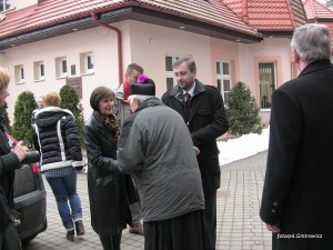 Osoby rozmawiają przed budynkiem z czerwonym dachem, na wyłożonym kostką brukowej podjeździe.