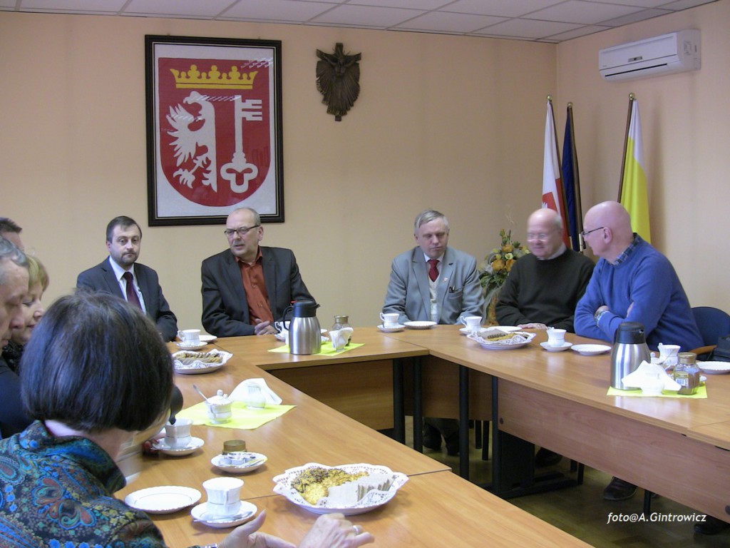 Grupa osób uczestniczy w spotkaniu przy stole w pomieszczeniu z herbem i flagami na ścianie.