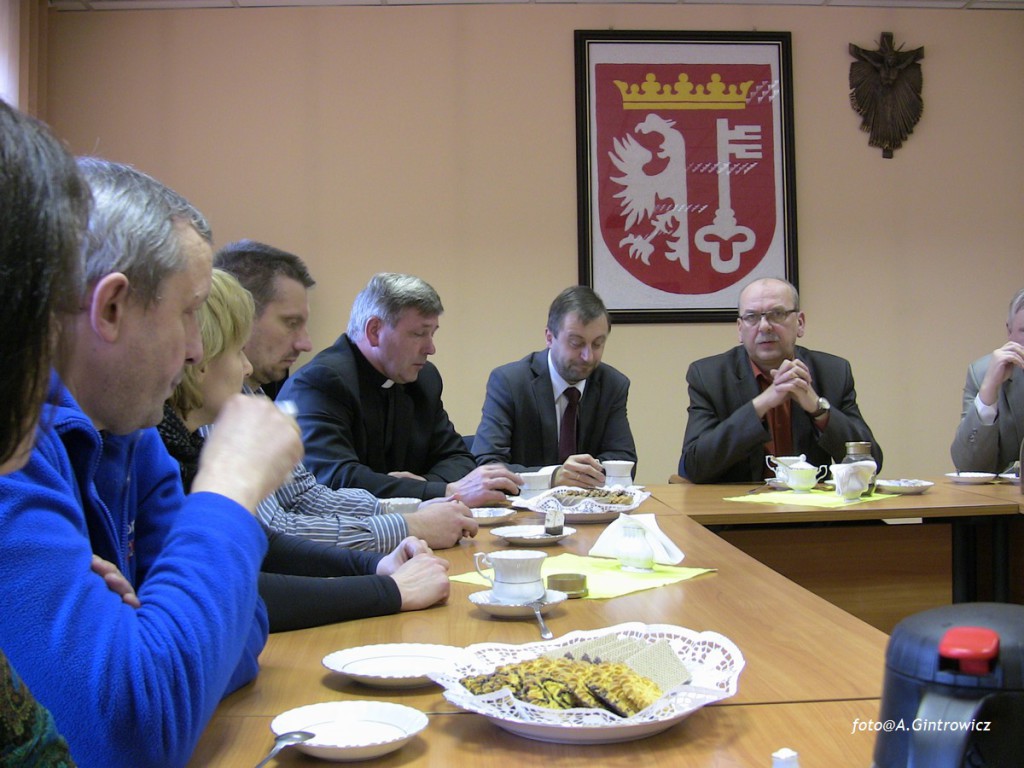 Uczestnicy spotkania przy stole, w tle widoczny herb oraz dekoracja ścienna.
