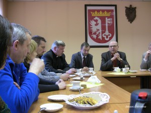 Uczestnicy spotkania przy stole, w tle widoczny herb oraz dekoracja ścienna.