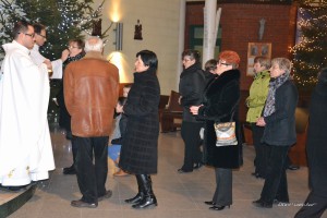 Uczestnicy ceremonii religijnej wewnątrz budynku, w tle widoczne dekoracje świąteczne i obrazy.