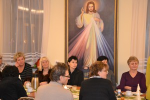 Grupa osób uczestniczy w spotkaniu przy stole, w tle widoczny obraz przedstawiający Jezusa Miłosiernego.