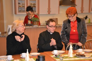 Grupa osób siedzi przy stole w pomieszczeniu, widoczne elementy wystroju i zastawa stołowa.