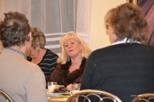 Grupa osób siedzi przy stole w jadalni, widoczne są talerze z jedzeniem i kubek.