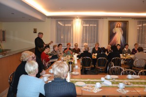Grupa osób uczestniczy w spotkaniu, podczas którego mężczyzna gra na gitarze przy długim stole nakrytym.