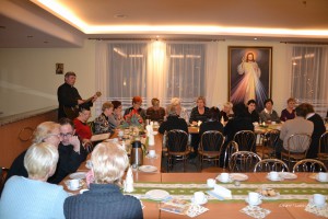 Grupa osób uczestniczy w spotkaniu, podczas którego muzyk gra na gitarze, a na ścianie wisi duży obraz.