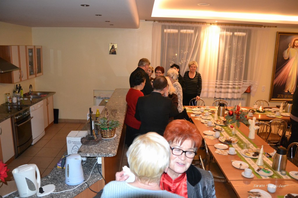 Wnętrze domu z grupą osób zgromadzonych przy długim stole nakrytym do posiłku.