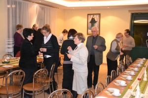 Grupa osób uczestniczy w spotkaniu przy długim stole nakrytym do posiłku w jasnym pomieszczeniu.