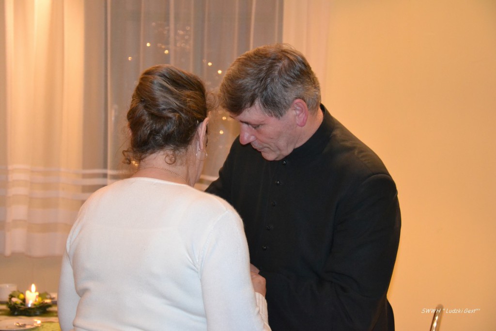 Dwie osoby stoją blisko siebie, patrząc w dół. W tle widoczne zasłony i dekoracje.