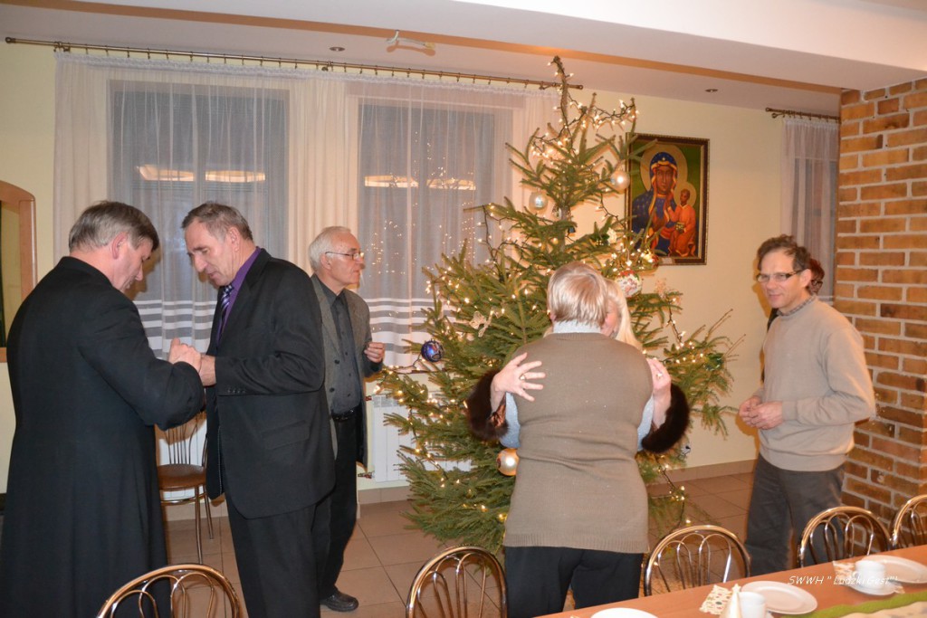 Uczestnicy spotkania w pomieszczeniu z choinką i obrazem religijnym, w tle widoczne okna i meble.