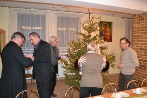 Uczestnicy spotkania w pomieszczeniu z choinką i obrazem religijnym, w tle widoczne okna i meble.