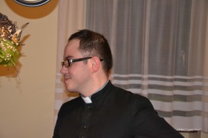 Mężczyzna w czarnym habitie stoi w pomieszczeniu z zasłonami i bukietem kwiatów w tle.