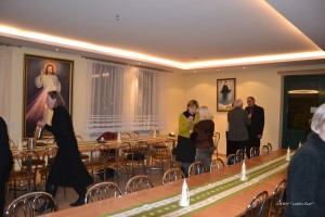 W sali znajdują się długie stoły z krzesłami, a w tle widać obrazy religijne i okna.