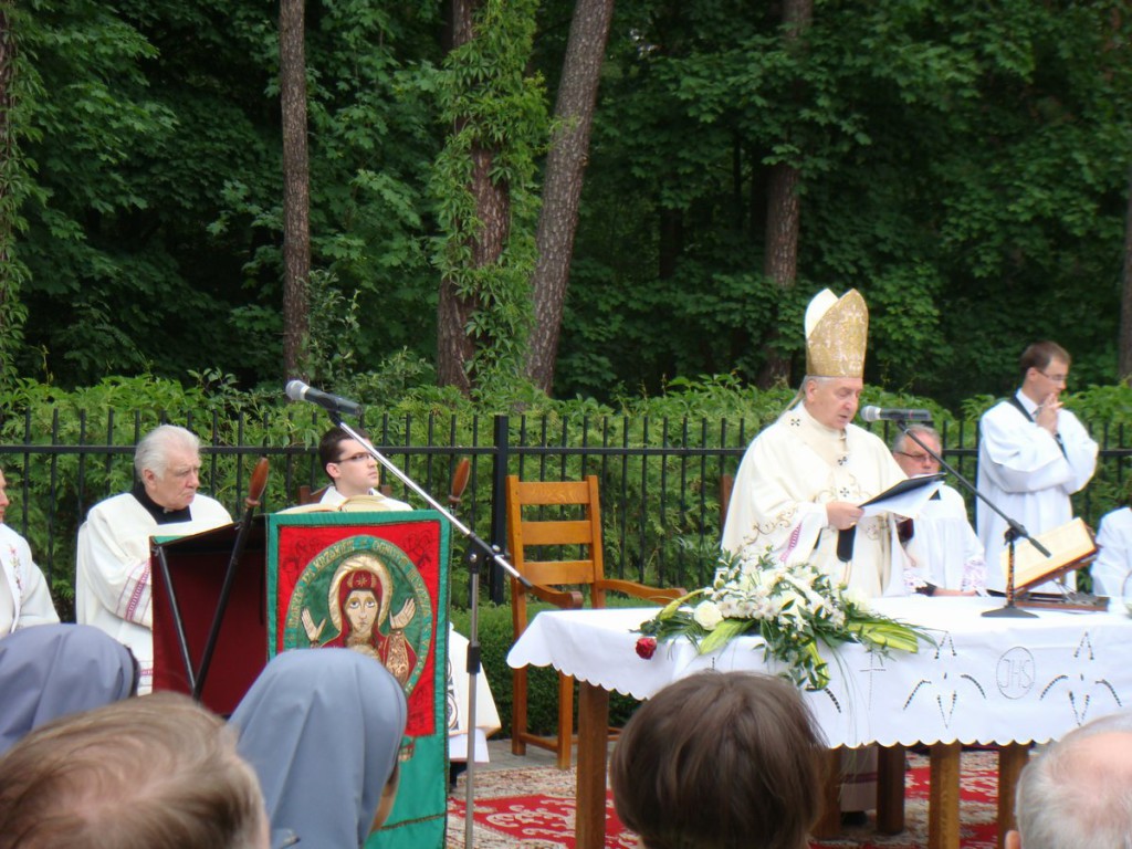 Uroczystość religijna odbywa się na zewnątrz, z udziałem duchownych i wiernych, w otoczeniu zieleni.