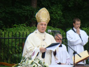 Biskup odprawia uroczystość w otoczeniu zieleni, w towarzystwie kapłanów i przy mikrofonie.