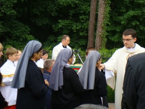 Uroczystość religijna odbywa się na zewnątrz, w otoczeniu zieleni. Obecni kapłan, wierni i siostry zakonne.