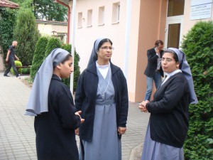 Trzy siostry zakonne rozmawiają na chodniku przed budynkiem z tablicą informacyjną „Hospicjum”.