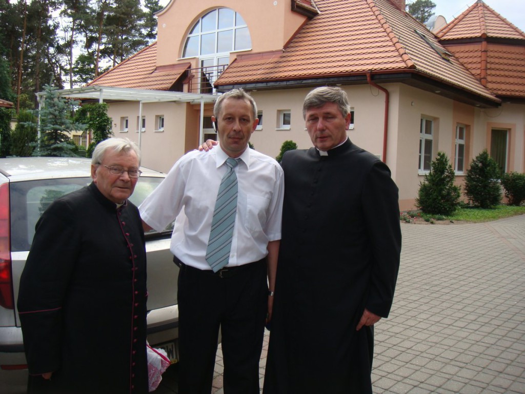 Trzej mężczyźni stoją przed budynkiem, dwóch w odzieży duchownej, jeden w stroju biznesowym.
