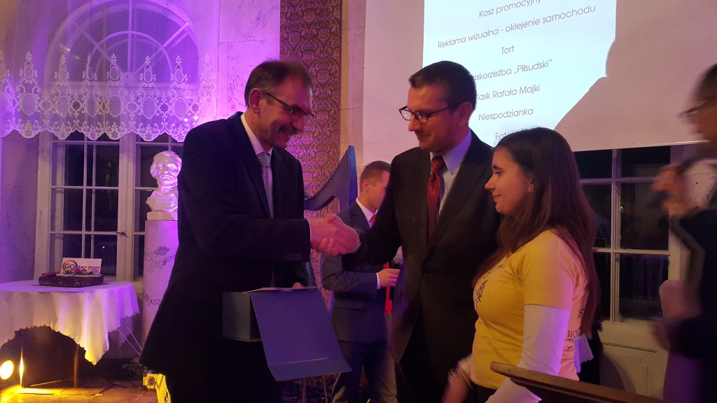 Dwie osoby w garniturach wręczają prezent osobie w żółtej bluzie, w tle widoczne dekoracje i projektor.