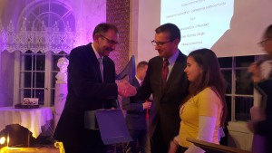 Dwie osoby w garniturach wręczają prezent osobie w żółtej bluzie, w tle widoczne dekoracje i projektor.