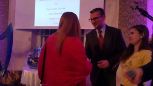Mężczyzna w garniturze rozmawia z dwiema osobami podczas toastu Noworocznego, w tle widoczny ekran z napisem.
