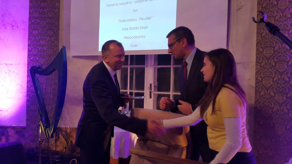 Mężczyźni w garniturach wręczają nagrodę osobie w żółtej bluzce, w tle widoczny projektor z prezentacją.