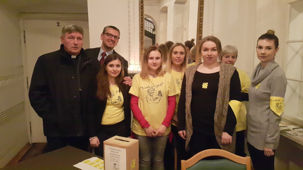 Grupa osób w żółtych koszulkach zbiera datki do puszki podczas imprezy Noworocznej w hospicjum w 2017 roku.