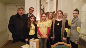 Grupa osób w żółtych koszulkach zbiera datki do puszki podczas imprezy Noworocznej w hospicjum w 2017 roku.