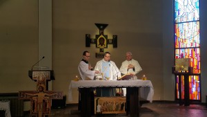 Trzej mężczyźni w szatach odprawiają liturgię przy ołtarzu w jasnym, bogato zdobionym wnętrzu kościoła.